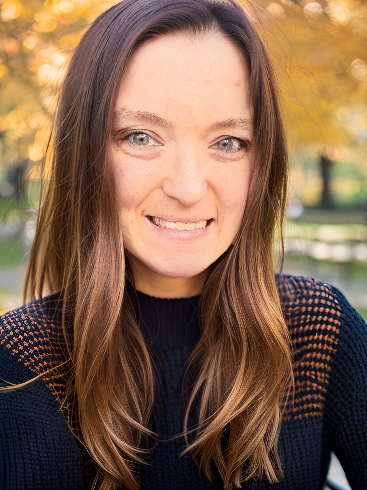 Person with long brown hair wearing a dark knitted sweater, outdoors with autumn leaves in the background.