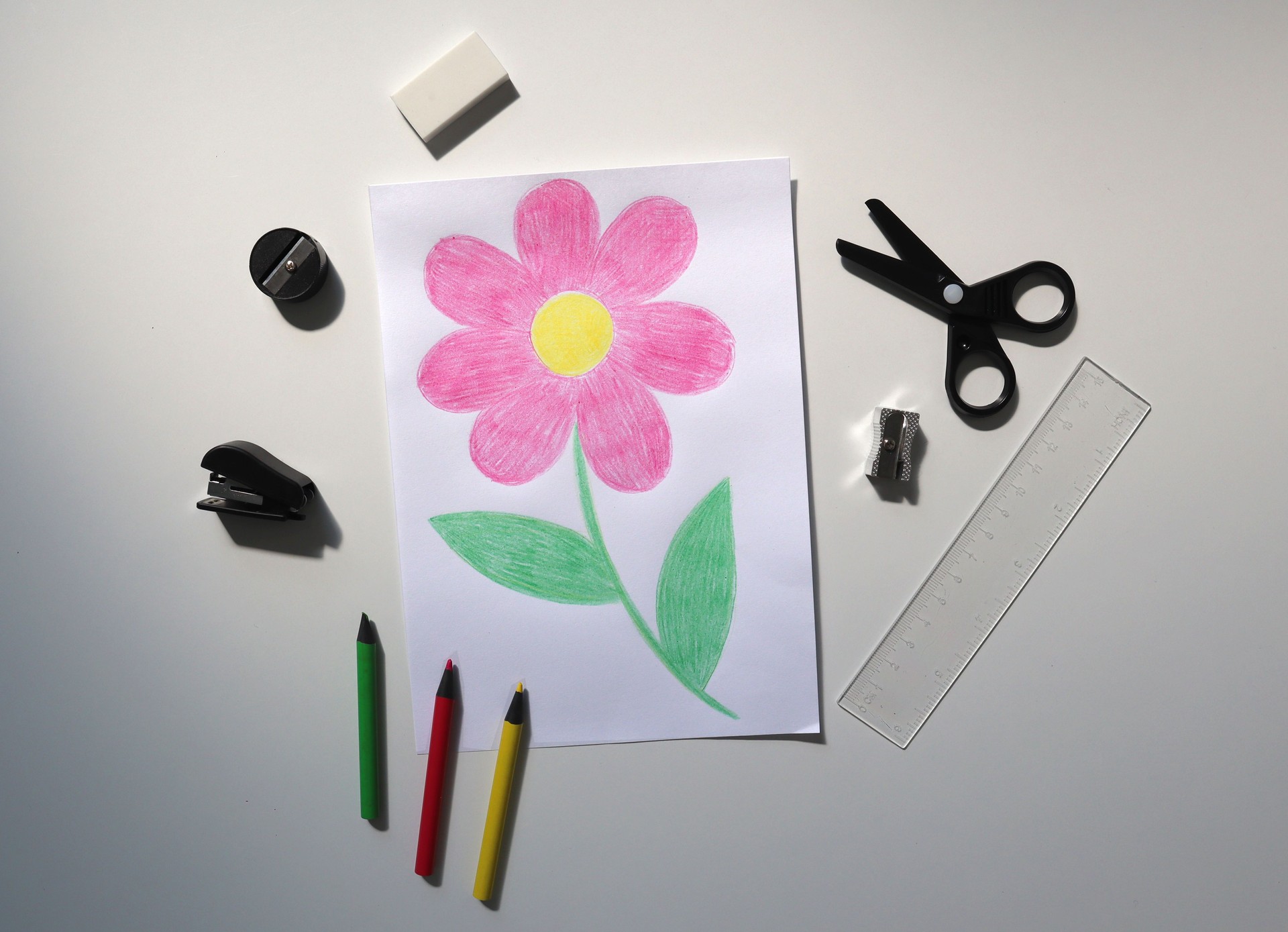 Photo of a child's drawing with colored pencils. Drawing of a flower. Pencils are nearby. White background.