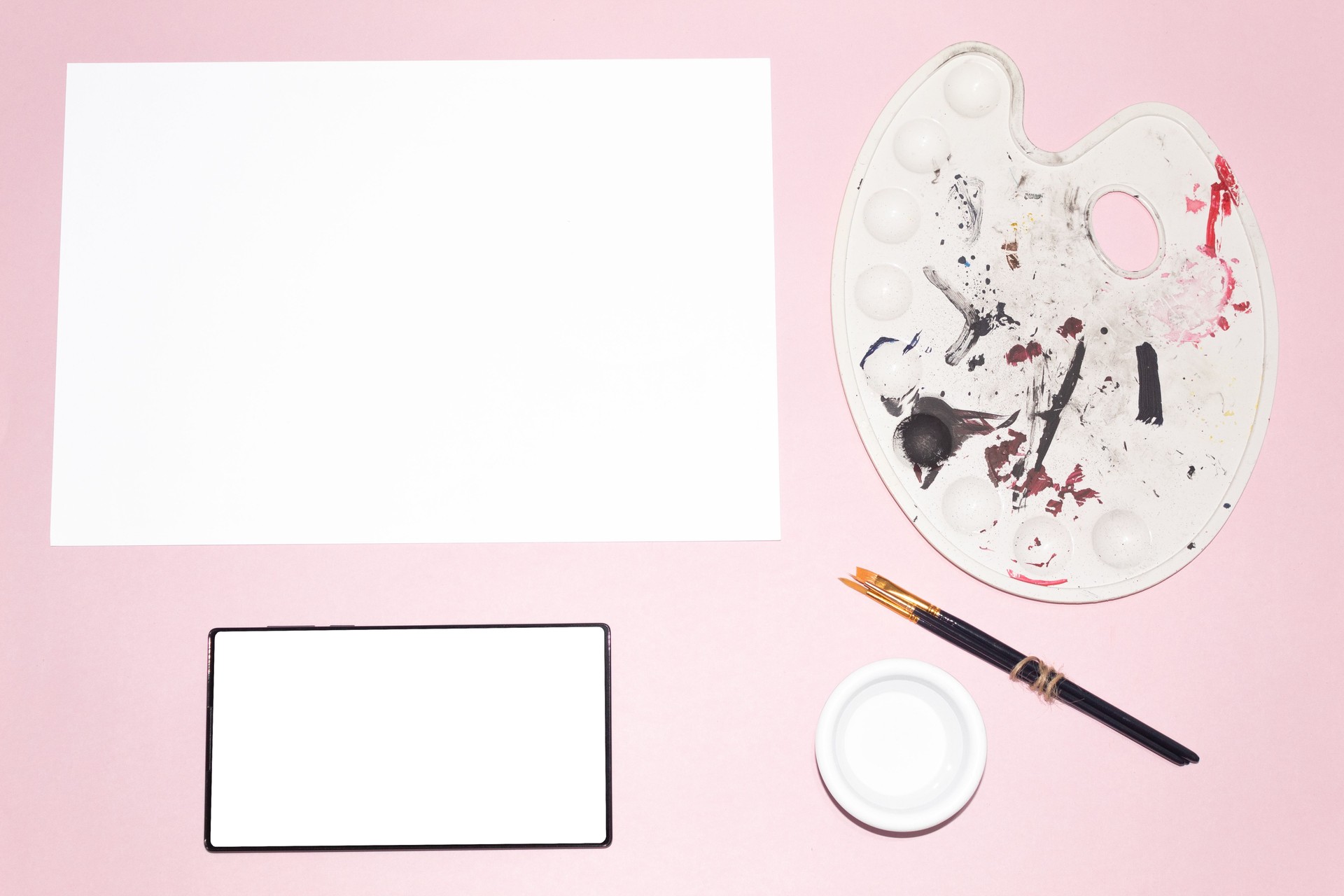 A white cardboard and a tablet with a blank screen next to a set of painting materials. Artistic mockup. Concept of technology and art.